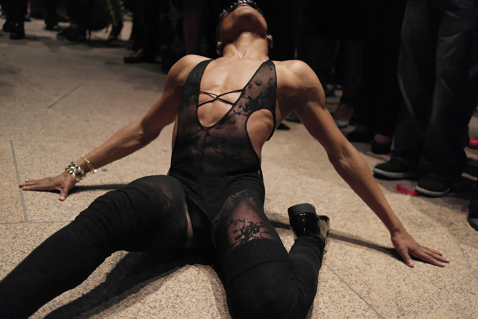 A person in an expressive dance pose wearing a black lace bodysuit and platform boots, performing on a stone floor during a queer creative showcase.