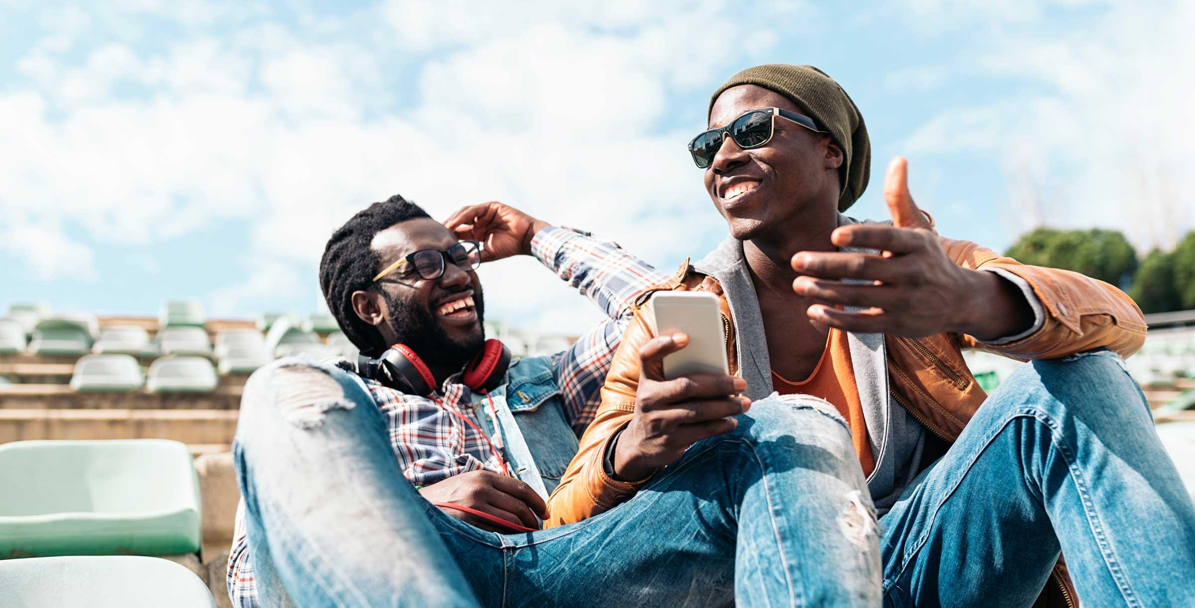 Two Black men laughing and sharing content on a smartphone outdoors, representing the Bessa community's focus on queer connection and found family.