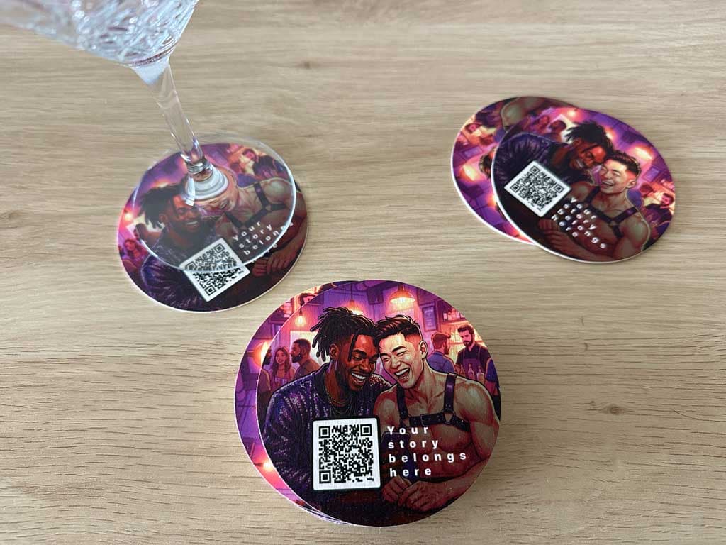 Free drink coasters for LGBT bars featuring a colorful illustration of two queer men smiling, a scannable QR code, and the text "Your story belongs here" on a wooden table.